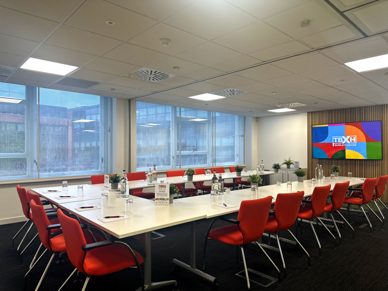 Boardroom-style conference room at TBXH with red chairs and a wall screen